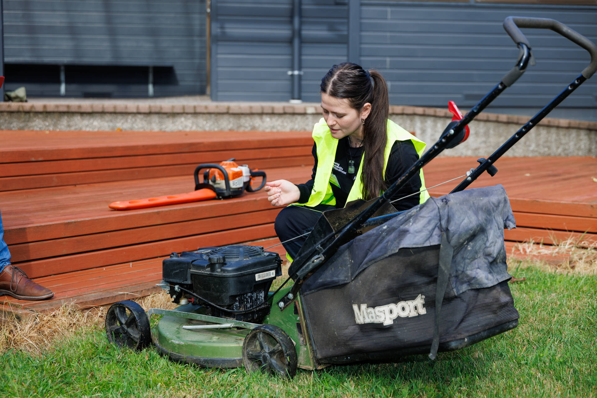 Person wearing a high-visibility vest kneeling on grass and inspecting a lawn mower, with gardening tools and a wooden deck in the background.