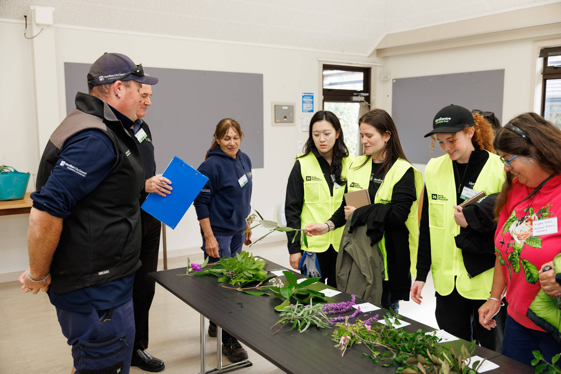 Group of people gathered around a table indoors examining and discussing different plant samples laid out in front of them, with several participants wearing high-visibility vests.