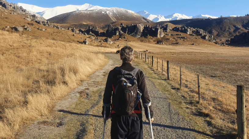 A man walks down a gravel road towards some hills, he is using crutches
