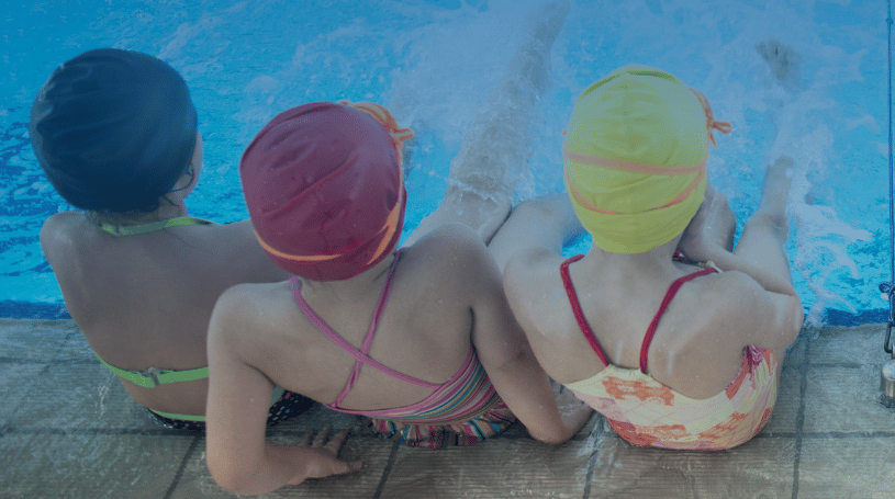 A group of children wearing brightly coloured swim caps and swimsuits sit on the edge of a swimming pool, their feet splashing in the water.