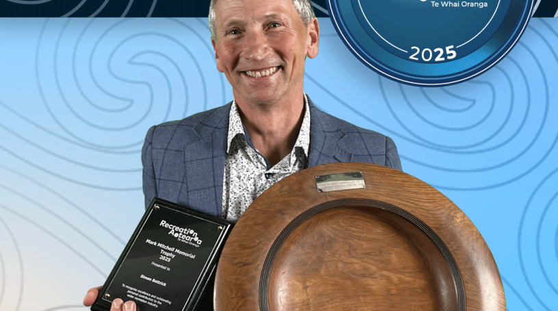 A man standing holding a large disc-shaped wooden award plate an award plaque while smiling. The background is a blue koru pattern with text at the top and bottom of the image. The top text says "2025 Recreation Awards Winner," and the bottom says "Mark Mitchell Memorial Trophy: Simon Battrick"