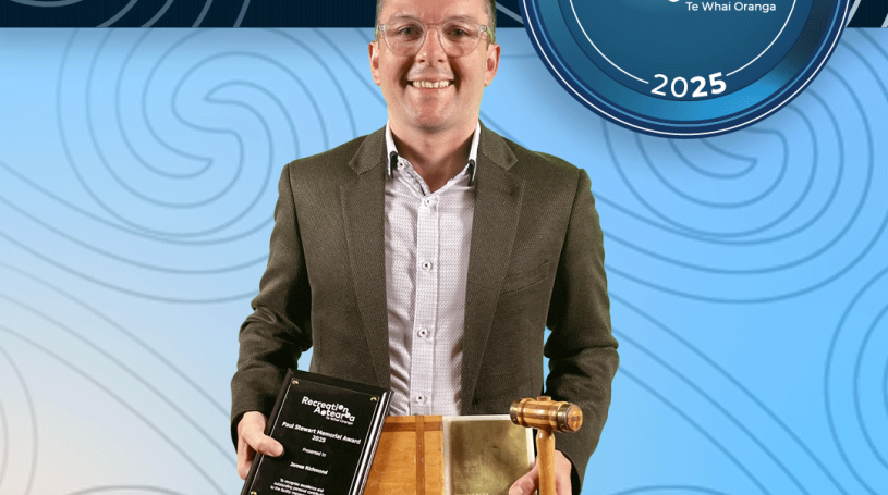 A man standing holding an award shaped like a gavel and sound block, as well as an award plaque and smiling. The background is a blue koru pattern with text at the top and bottom of the image. The top text says "2025 Recreation Awards Winner," and the bottom says "Paul Stuart Memorial Award: James Richmond"