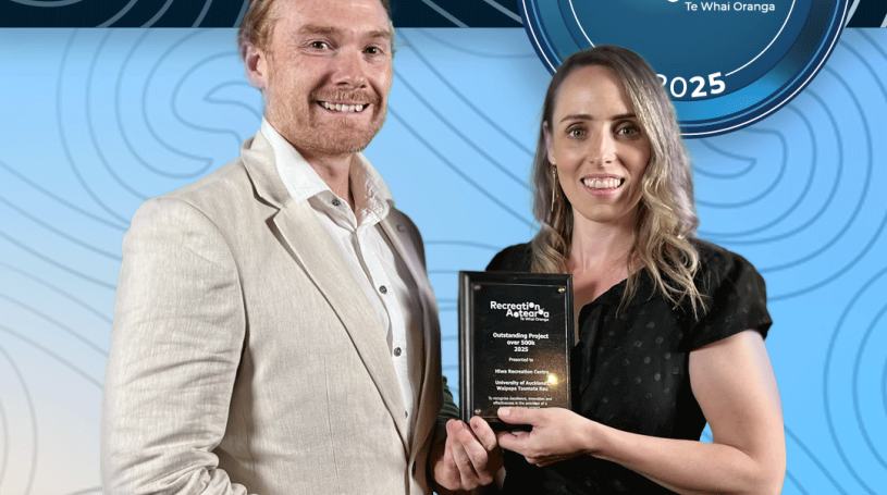 A man and woman standing together holding an award plaque and smiling. The background is a blue koru pattern with text at the top and bottom of the image. The top text says "2025 Recreation Awards Winner," and the bottom says "Outstanding Project over $500k: Hiwa Recreation Centre"