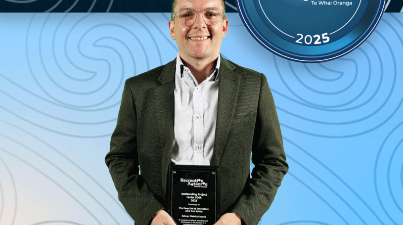 A man standing holding an award plaque and smiling. The background is a blue koru pattern with text at the top and bottom of the image. The top text says "2025 Recreation Awards Winner," and the bottom says "Outstanding Project under $500k: The Deep End of Innovation: AI and Pool Safety"