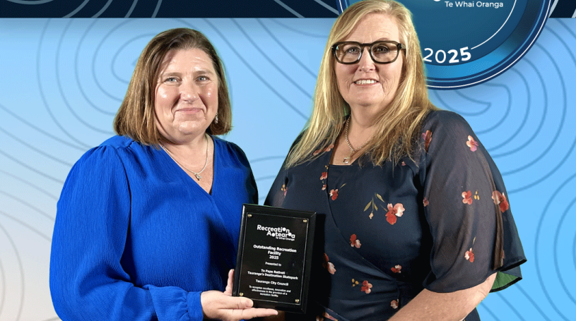 Two women standing together holding an award plaque and smiling. The background is a blue koru pattern with text at the top and bottom of the image. The top text says "2025 Recreation Awards Winner," and the bottom says "Outstanding Recreation Facility: Te Papa Retireti: Tauranga's Destination Skatepark"