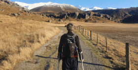 A man walks down a gravel road towards some hills, he is using crutches 