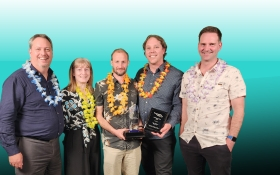 Five people stand smiling at a camera, two of them are holding awards. There is one lady and four men. They're all wearing leis.