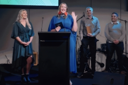 Three people stand on a stage. One is accepting an award and addressing the crowd.