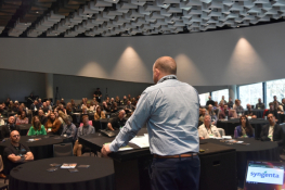 A man stands on a stage delivering a speech to the crowd. Photo is taken from behind him.