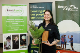 Person holding a wrapped plant and a wooden trophy plaque while standing indoors in front of banners for Horticentre Charitable Trust and Recreation Aotearoa.