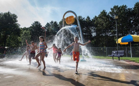 A group of children playing in togs at an outdoor splash pad. They are running towards the camera and away from a tall bucket fountain which has just tipped over, splashing water over them.