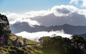 Four people cycle down a mountain bike track. In the background is hills that are covered in clouds
