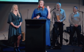 Three people stand on a stage. One is accepting an award and addressing the crowd.