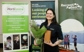 Person holding a wrapped plant and a wooden trophy plaque while standing indoors in front of banners for Horticentre Charitable Trust and Recreation Aotearoa.