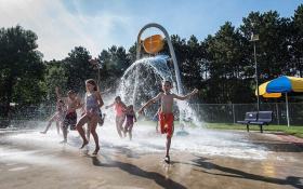 A group of children playing in togs at an outdoor splash pad. They are running towards the camera and away from a tall bucket fountain which has just tipped over, splashing water over them.